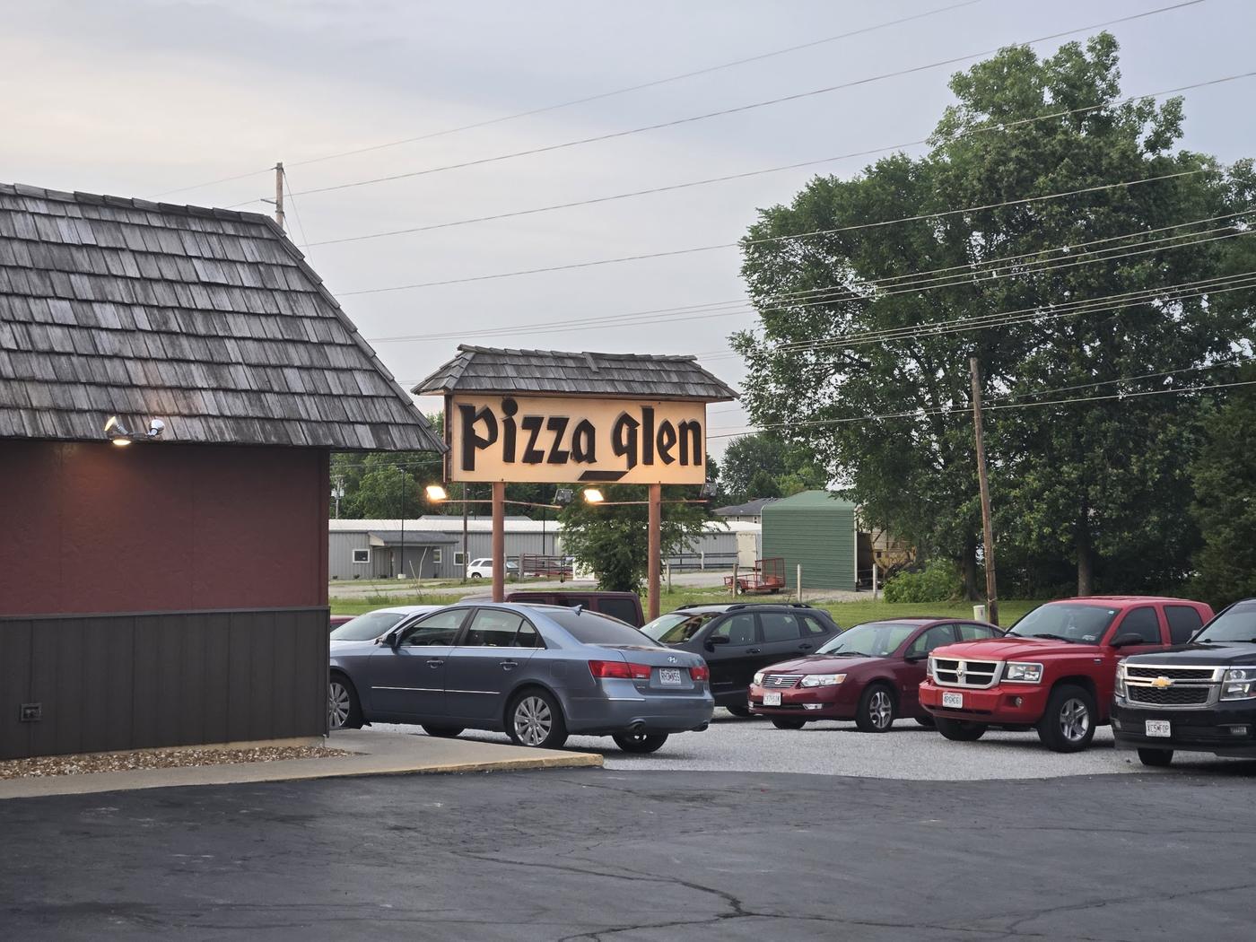 The lit Pizza Glen roadside sign at dusk, capped with its own small cedar shingle roof, with a full parking lot behind it